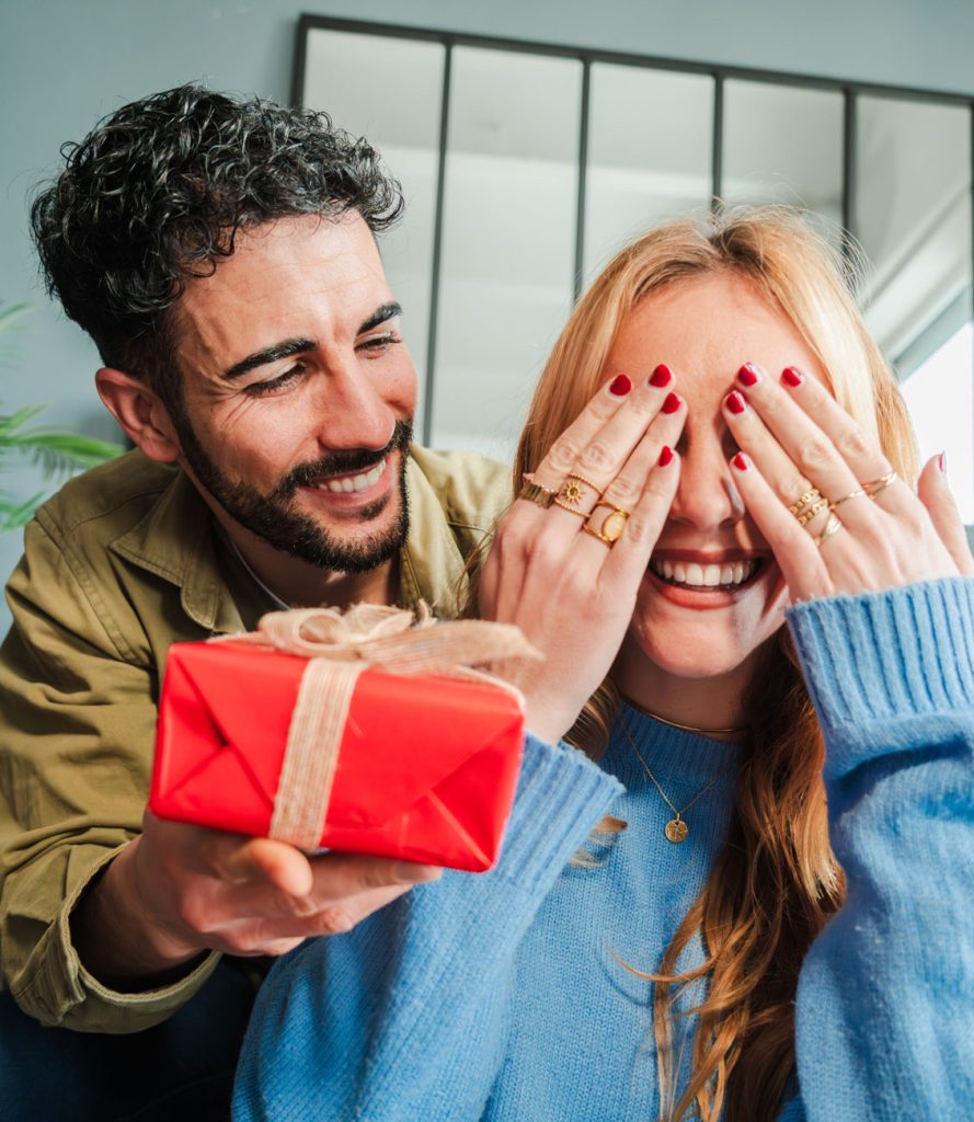 Un homme tend un cadeau rouge à une femme qui a les mains devant les yeux.